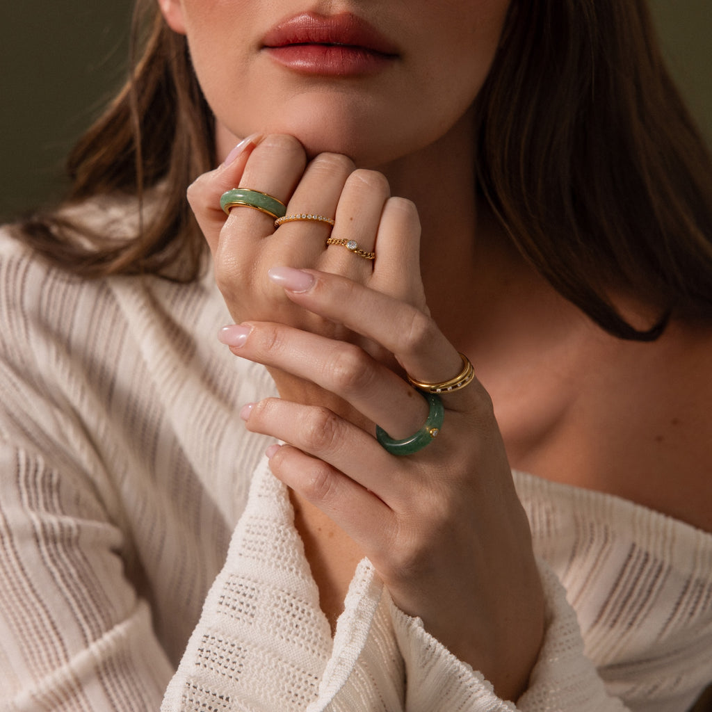A woman in a cream sweater displays her hands, one adorned with the gleaming Haven Jade Spinner Ring, gold band, and green bangles as her lips rest softly parted.