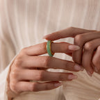 A hand with long, manicured nails holds the Haven Jade Spinner Ring—an elegant jade and gold band—against a cream textured background for a mindful jewelry look.