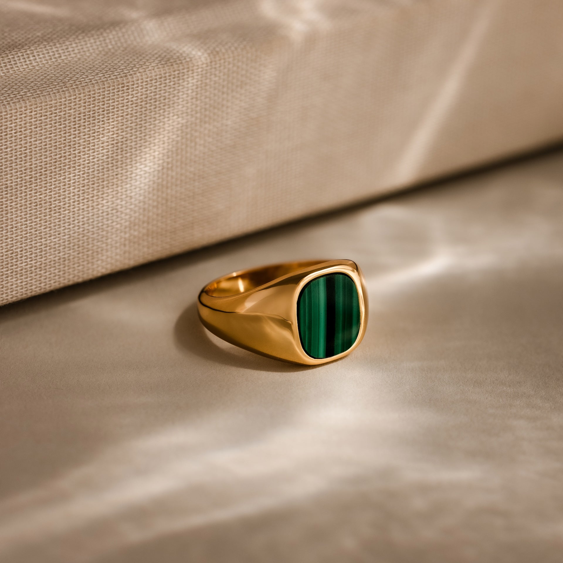 The Malachite Signet Ring, featuring a rectangular green stone symbolizing transformation, is displayed on a beige textured surface with soft light reflections.
