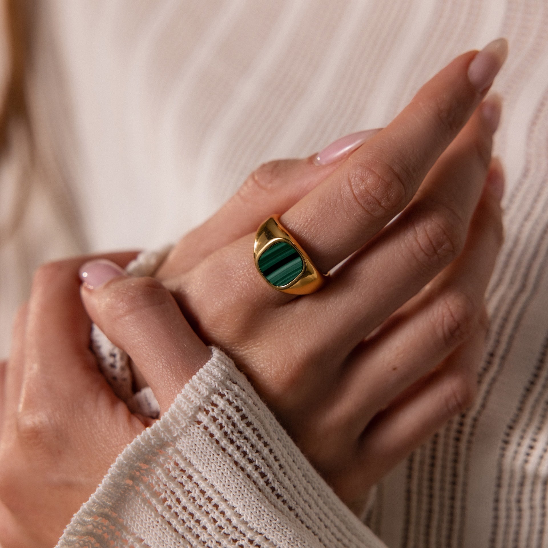 A hand with manicured nails wears the Malachite Signet Ring, featuring the transformative green stone, against a textured white sleeve.