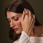 A woman with long brown hair touches her face, wearing a cream knit top and the Malachite Signet Ring—a striking green stone accessory symbolizing transformation.