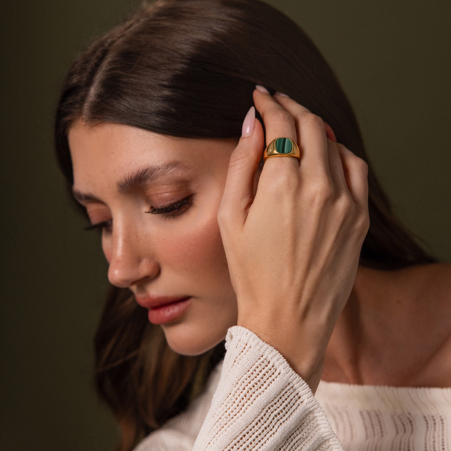 A woman with long brown hair touches her face, wearing a cream knit top and the Malachite Signet Ring—a striking green stone accessory symbolizing transformation.