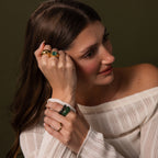 Woman with long brown hair wears gold rings, a Malachite Signet Ring—the stone of transformation—and a green bracelet, styled in a white off-shoulder sweater.