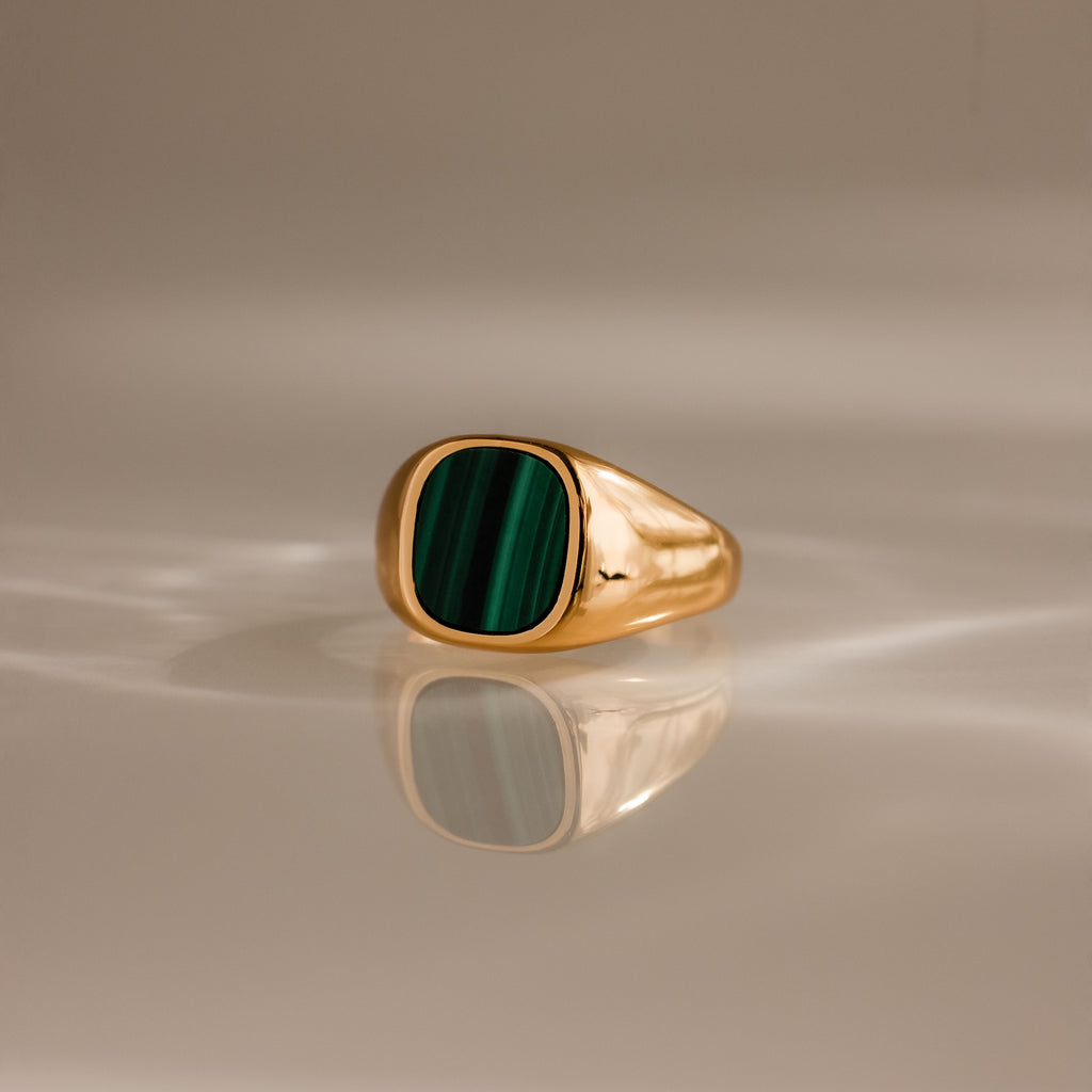 The Malachite Signet Ring features a rectangular green malachite stone set in gold, displayed on a reflective, softly lit surface.