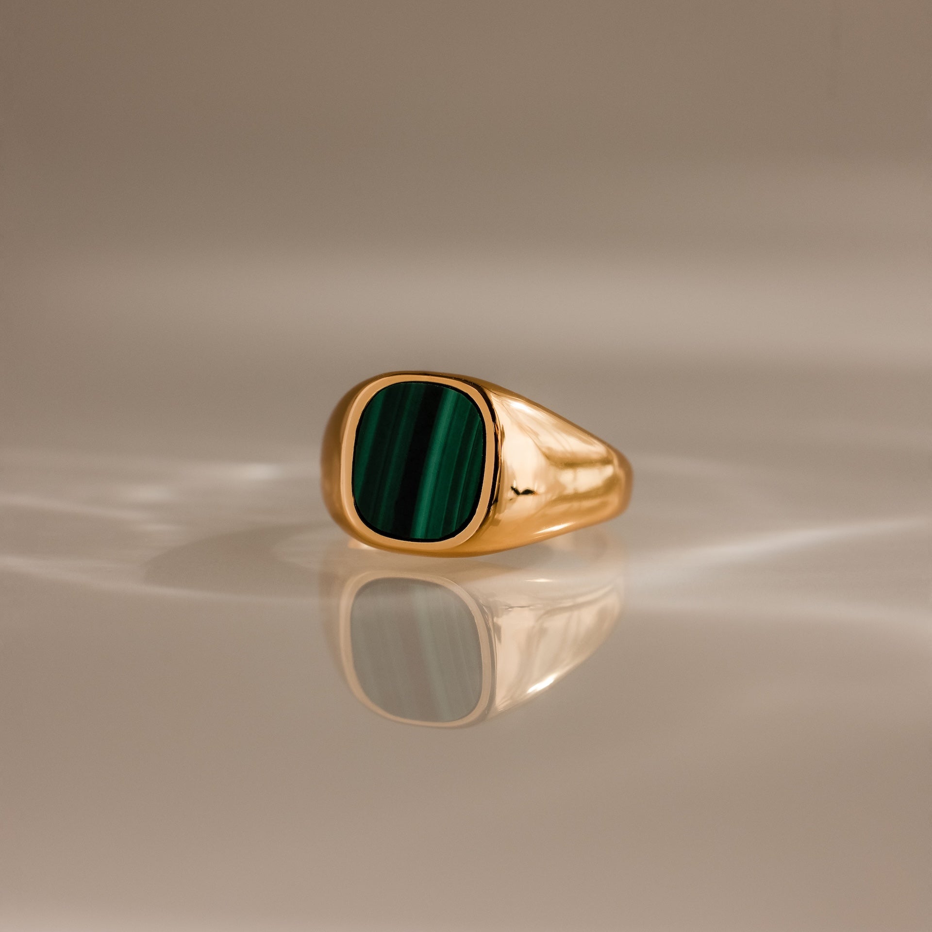 The Malachite Signet Ring features a rectangular green malachite stone set in gold, displayed on a reflective, softly lit surface.