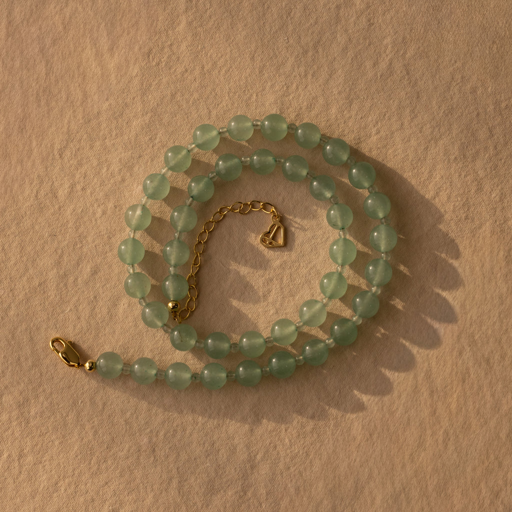 The Jade Choker Necklace in 18K Gold features green jade beads, a heart charm, and a gold clasp, elegantly coiled on a textured beige surface.