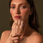 Woman with long brown hair wears the Skinny Lavender Jade Ring and gold rings, gazing calmly at the camera with her hand resting on her chin.