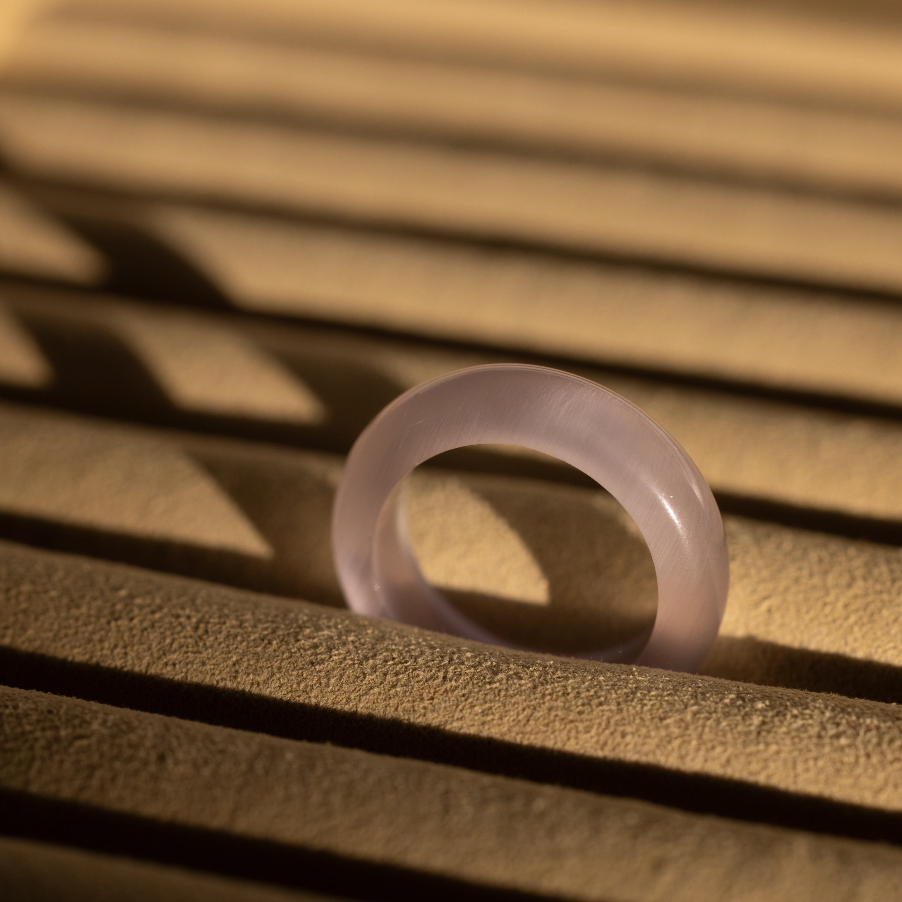 A Skinny Lavender Jade Ring rests on sand, its soothing energy mirrored in the dramatic shadows that stretch across the scene.