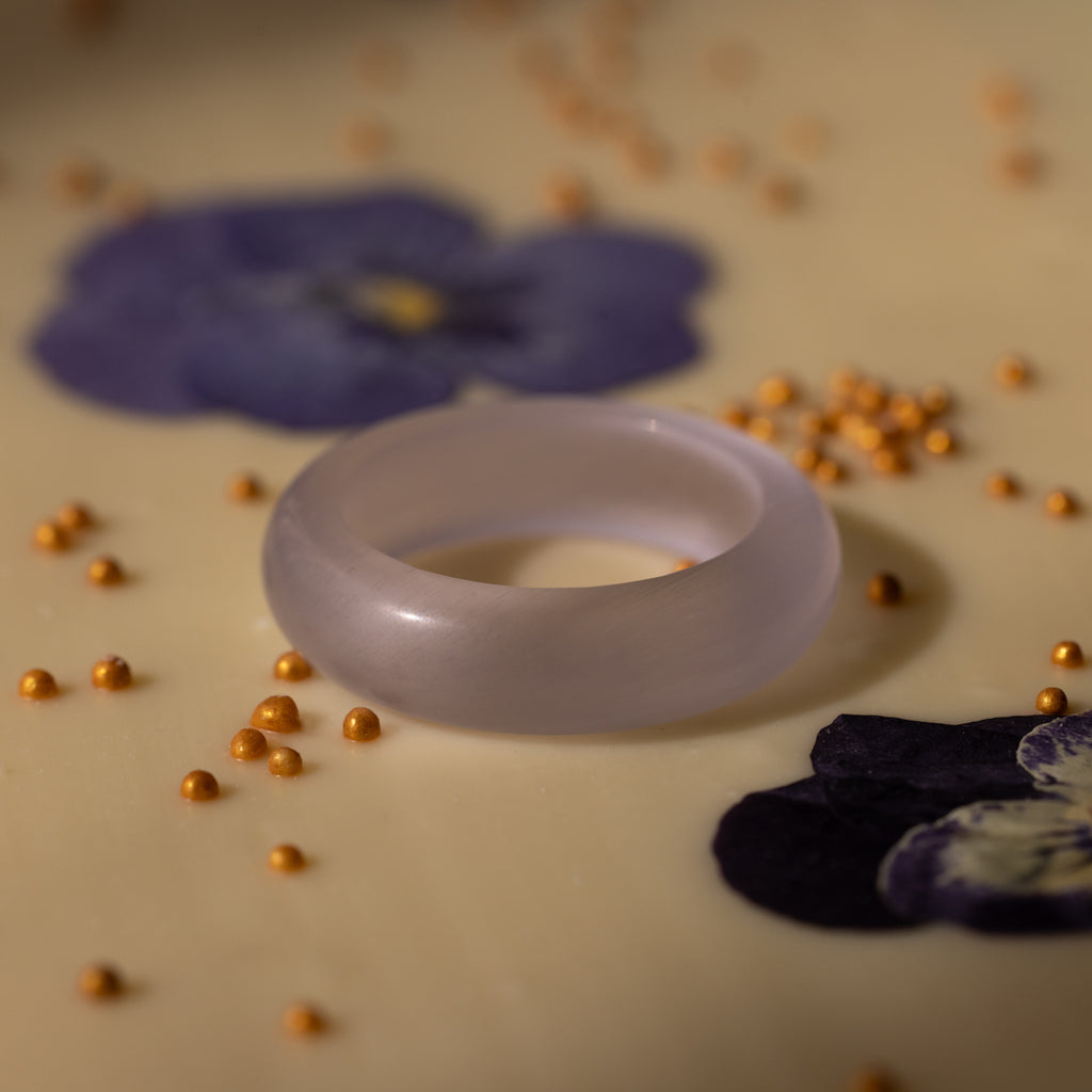 The Skinny Lavender Jade Ring rests on a surface with purple flowers and scattered yellow beads, radiating a calming energy.