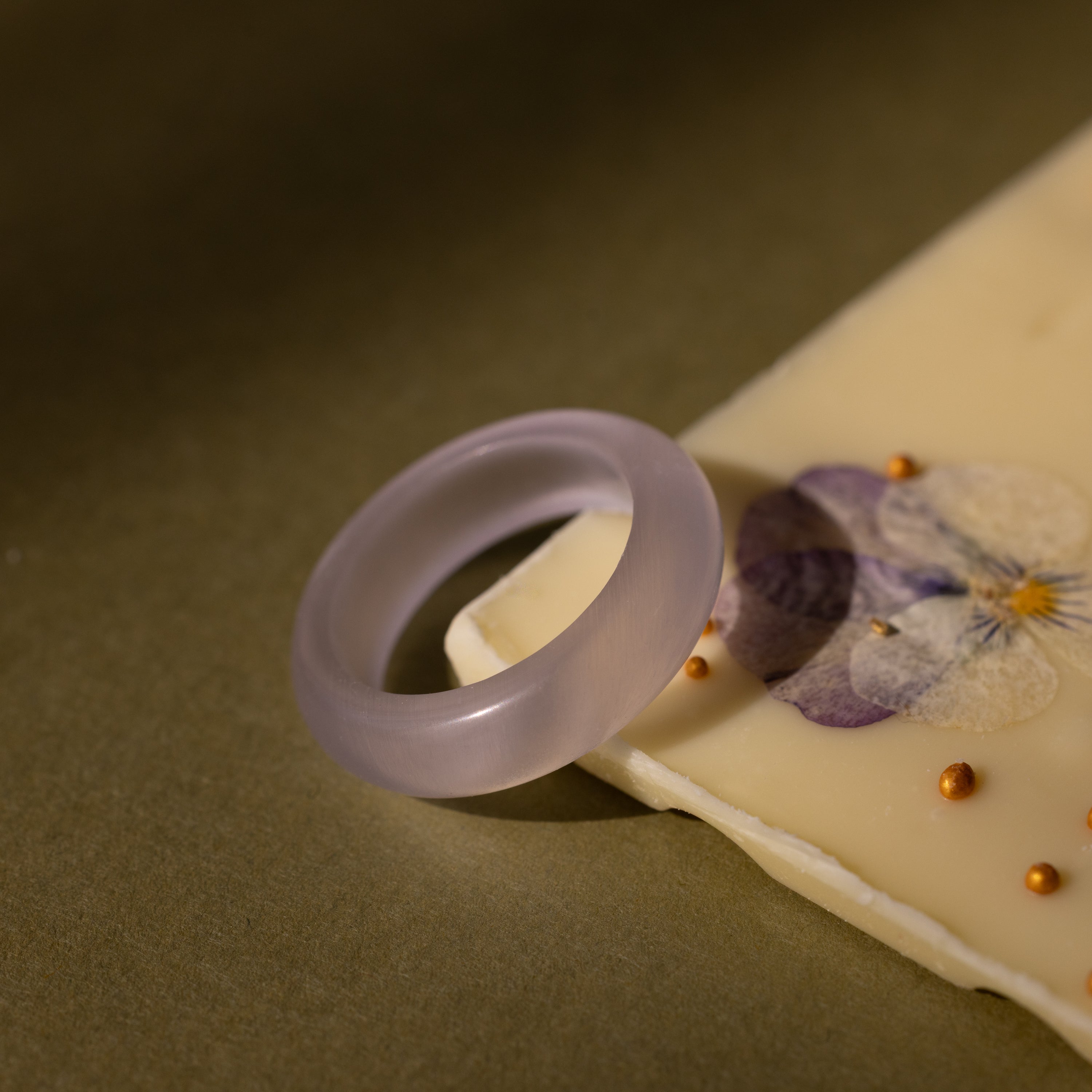 The Skinny Lavender Jade Ring, prized for its calming energy, sits atop a white chocolate bar embellished with dried flowers and gold beads.