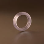 The Skinny Lavender Jade Ring, a translucent pale purple stacking ring, stands upright on a smooth brown surface under soft lighting, radiating calming energy.
