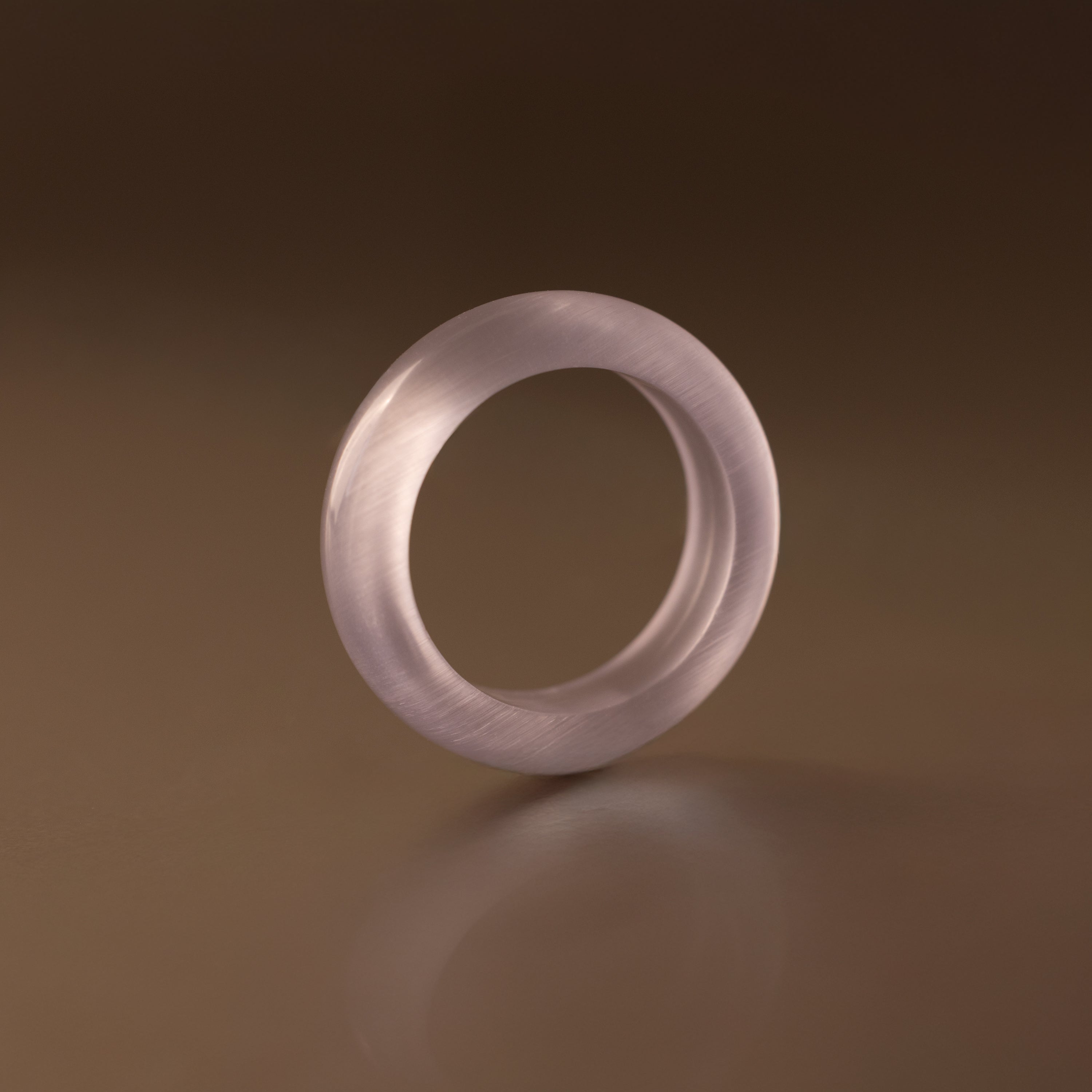 The Skinny Lavender Jade Ring, a translucent pale purple stacking ring, stands upright on a smooth brown surface under soft lighting, radiating calming energy.