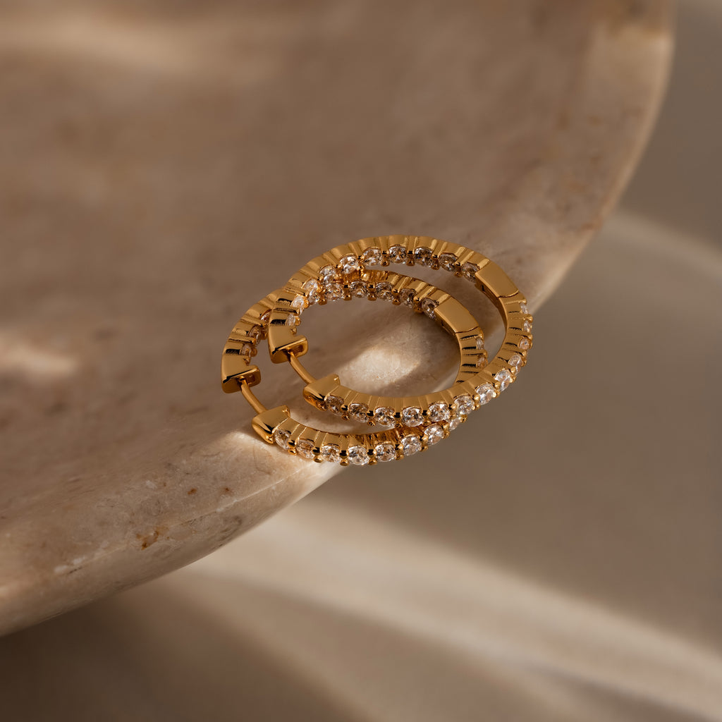 The Inside Out Diamond Hoops in 18K Gold, featuring clear stones in a classic huggie design, are displayed elegantly on a smooth beige marble surface.