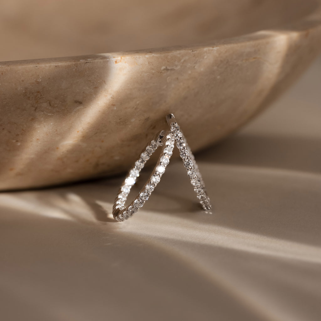 A pair of Inside Out Diamond Hoops in Silver rests against a beige marble bowl, illuminated by soft natural light.