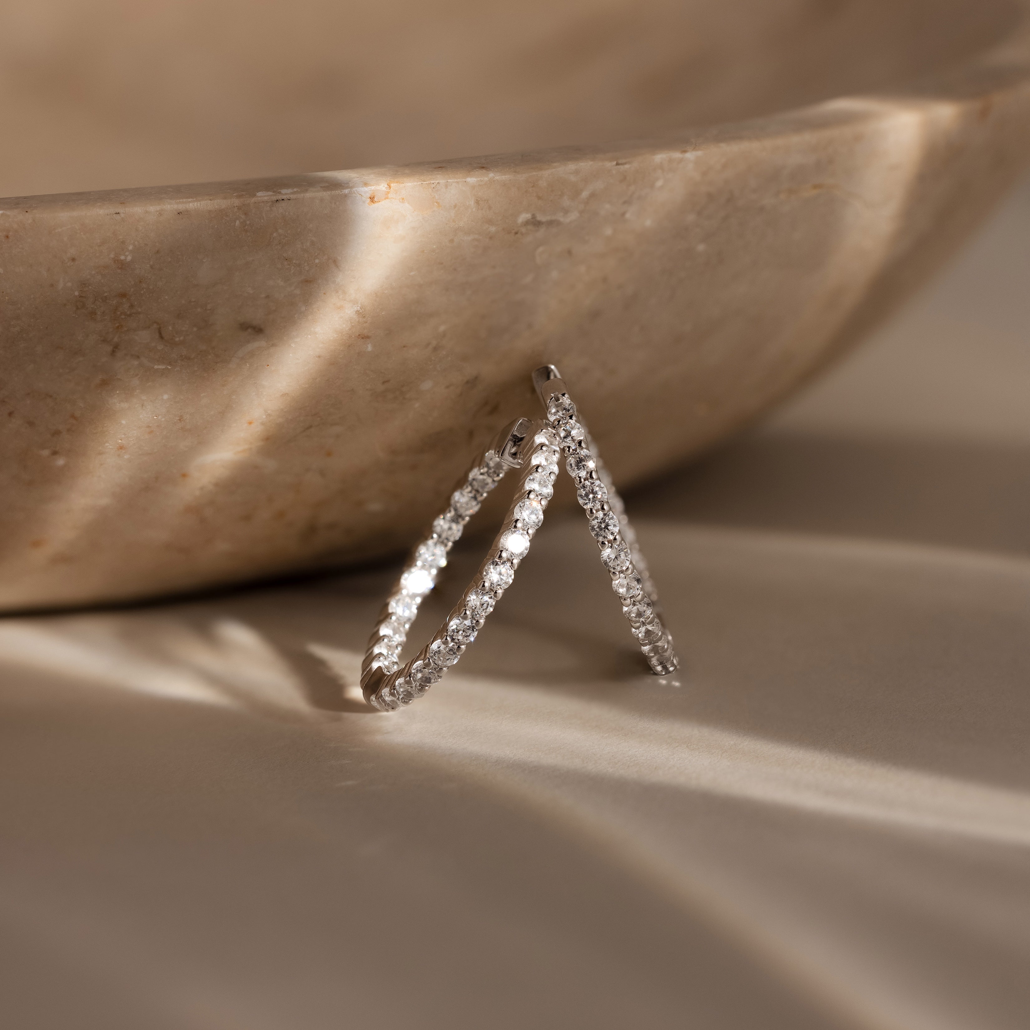 A pair of Inside Out Diamond Hoops in Silver rests against a beige marble bowl, illuminated by soft natural light.