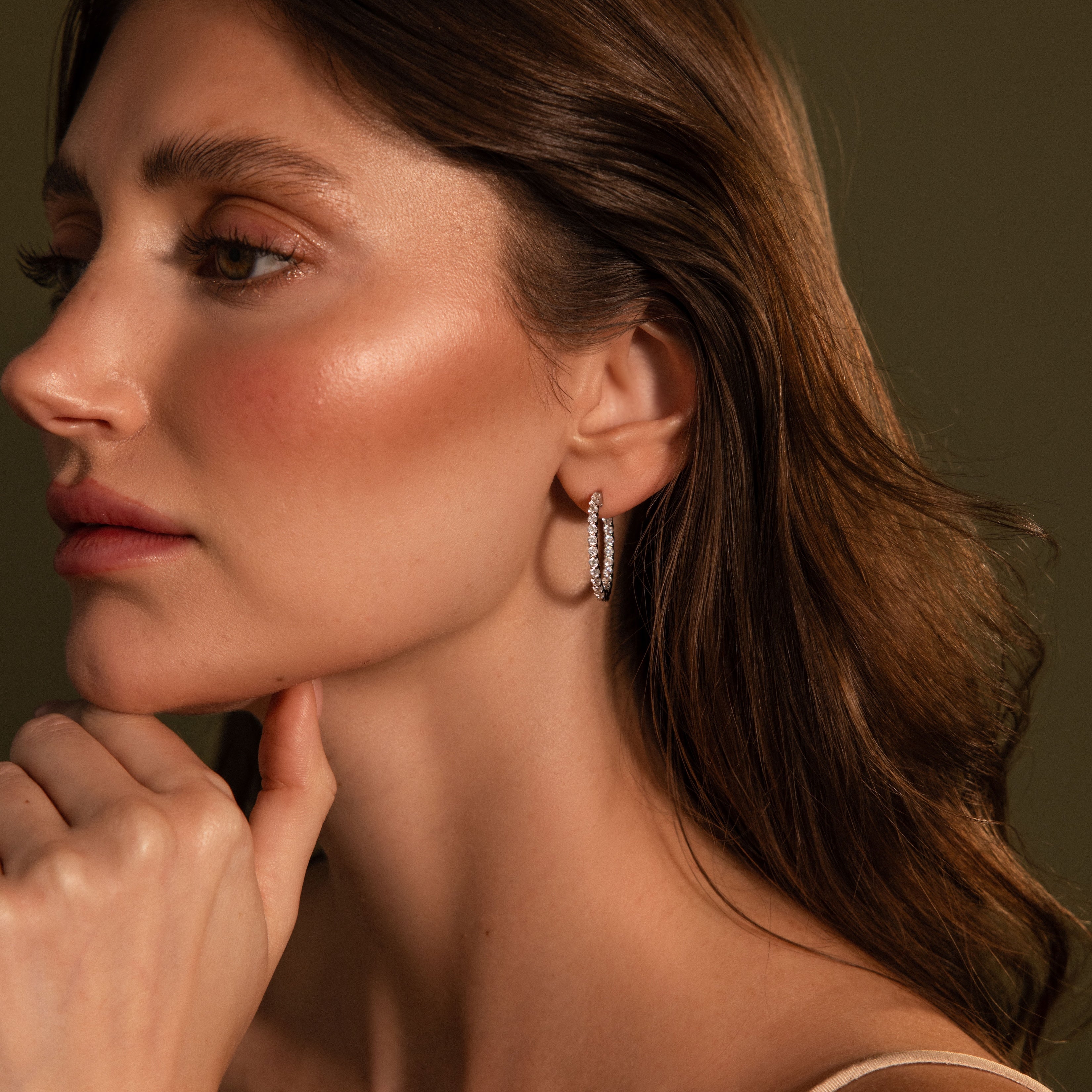 A woman with wavy brown hair wears Inside Out Diamond Hoops in Silver, gazing sideways with her hand near her chin against a dark background.