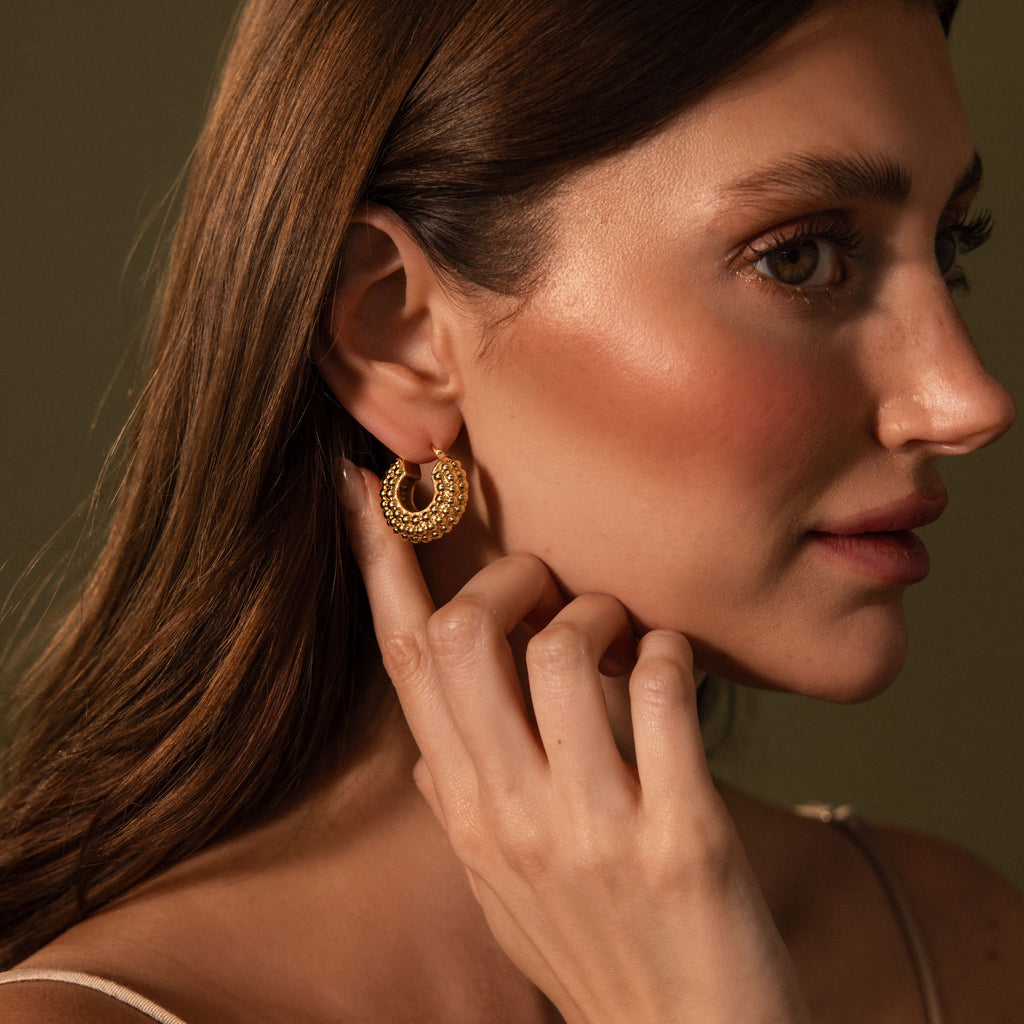 A woman with long brown hair touches Beaded Dome Hoops featuring gold beading against a neutral background.