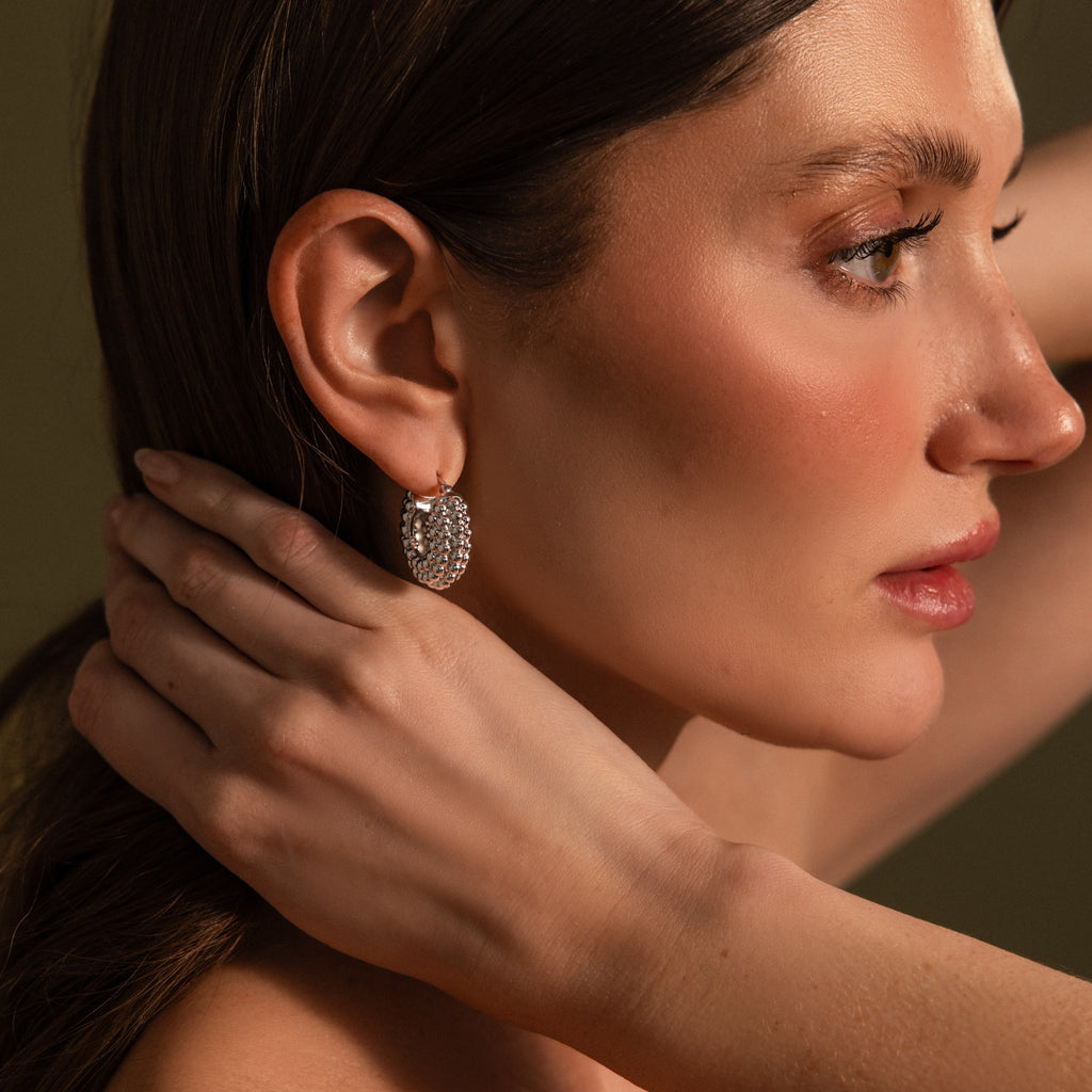 A woman with smooth skin wears Beaded Dome Hoops in Silver, featuring sparkling beads, as she touches her neck and gazes to the side.