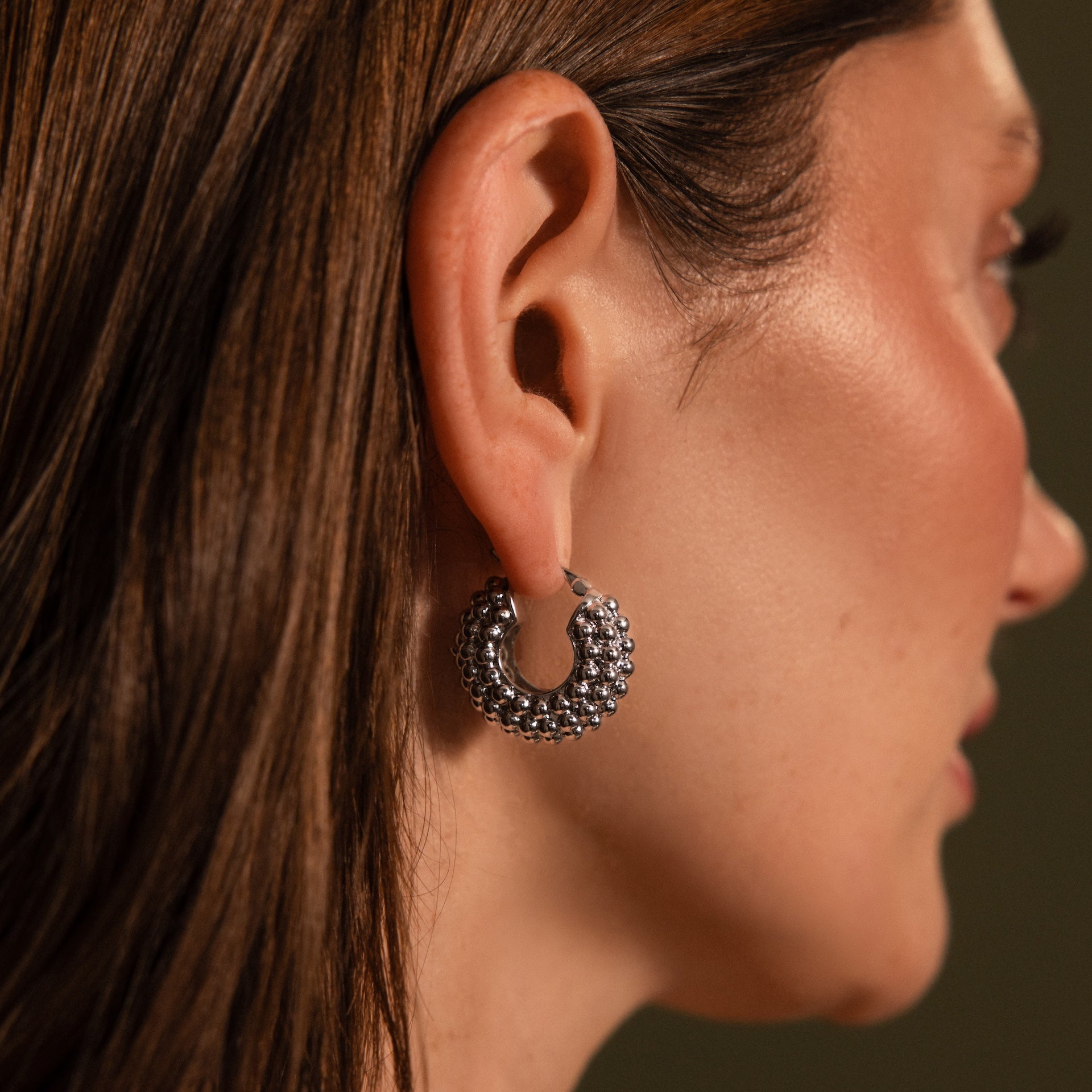 A woman with straight brown hair wears Beaded Dome Hoops in Silver on her left ear, showcasing statement silver hoops adorned with subtle gold beading.