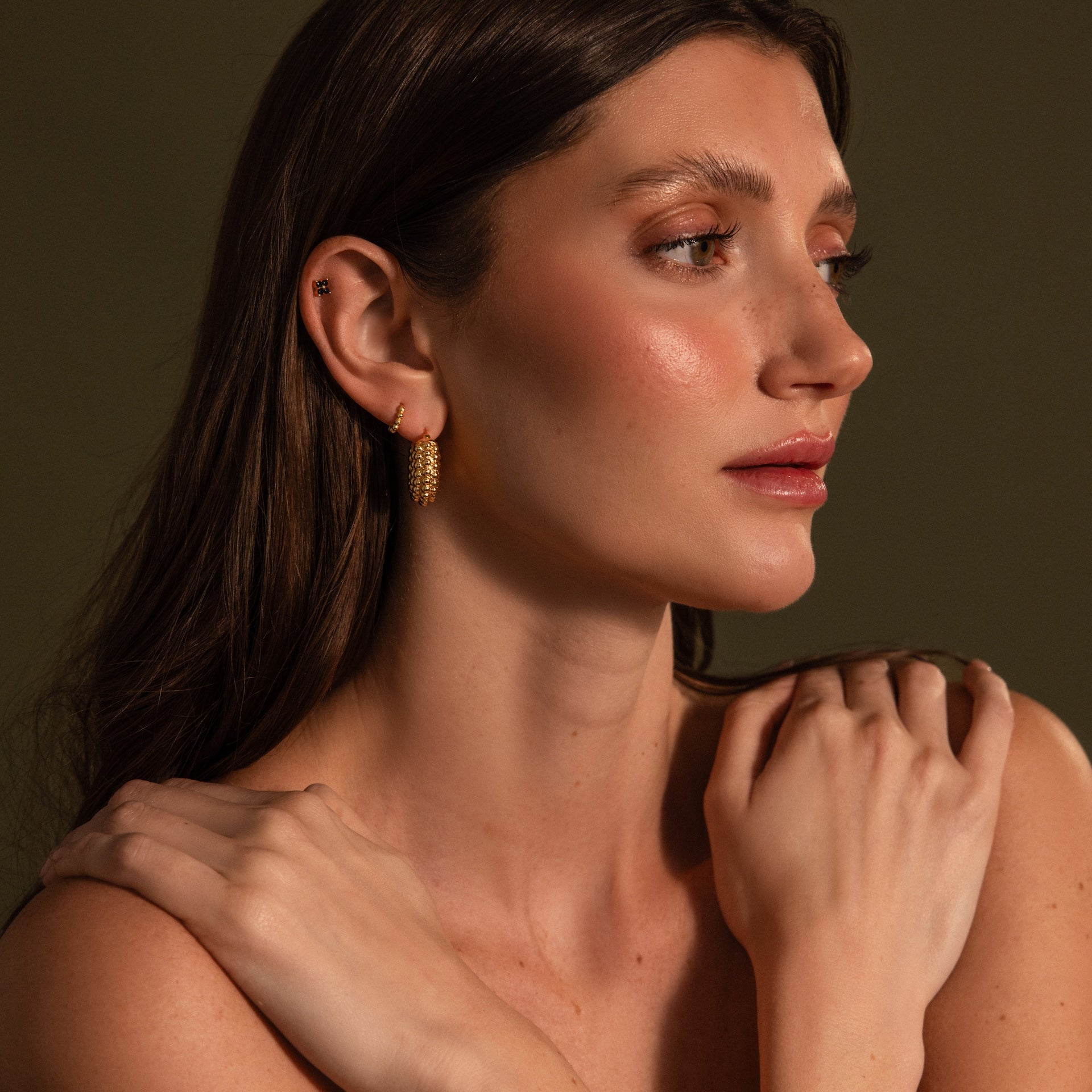 Woman with long brown hair wears Beaded Dome Hoops in 18K Gold, looking to the side with her hands resting on her shoulders.
