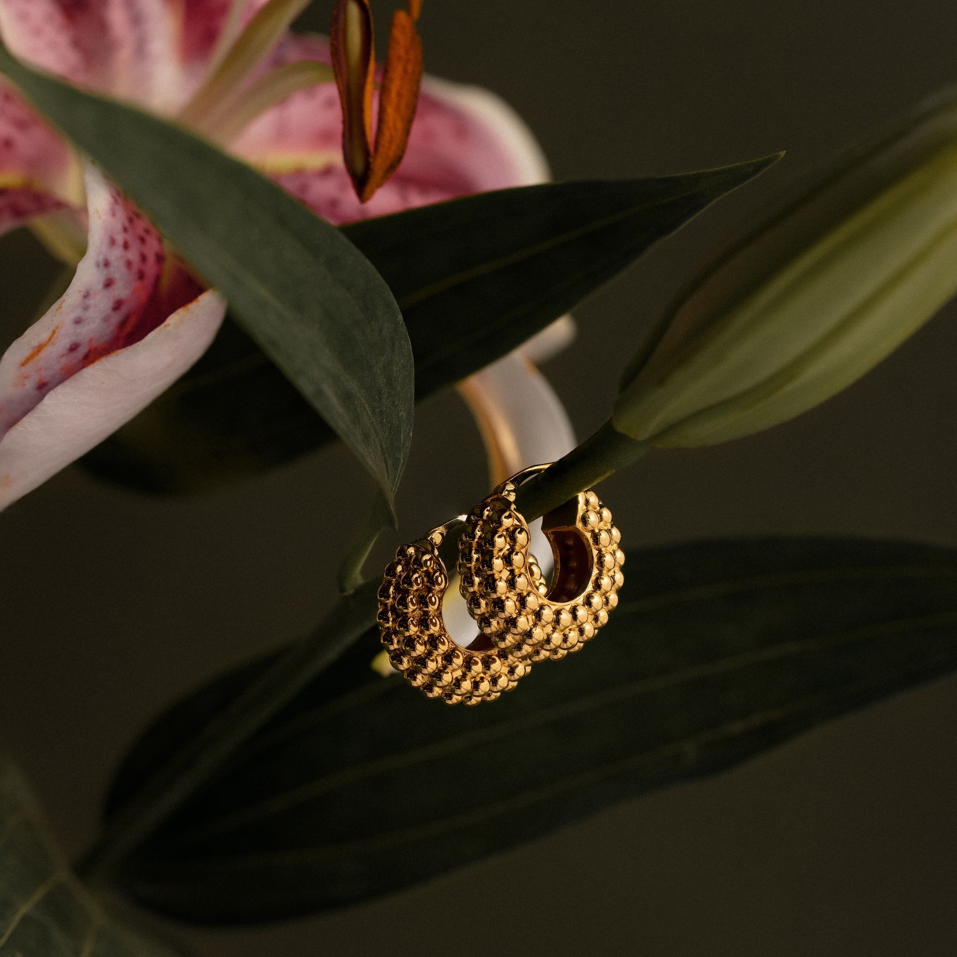 Beaded Dome Hoops in 18K Gold hang on a green leaf next to a pink and white lily flower, showcasing their elegant statement hoop design.