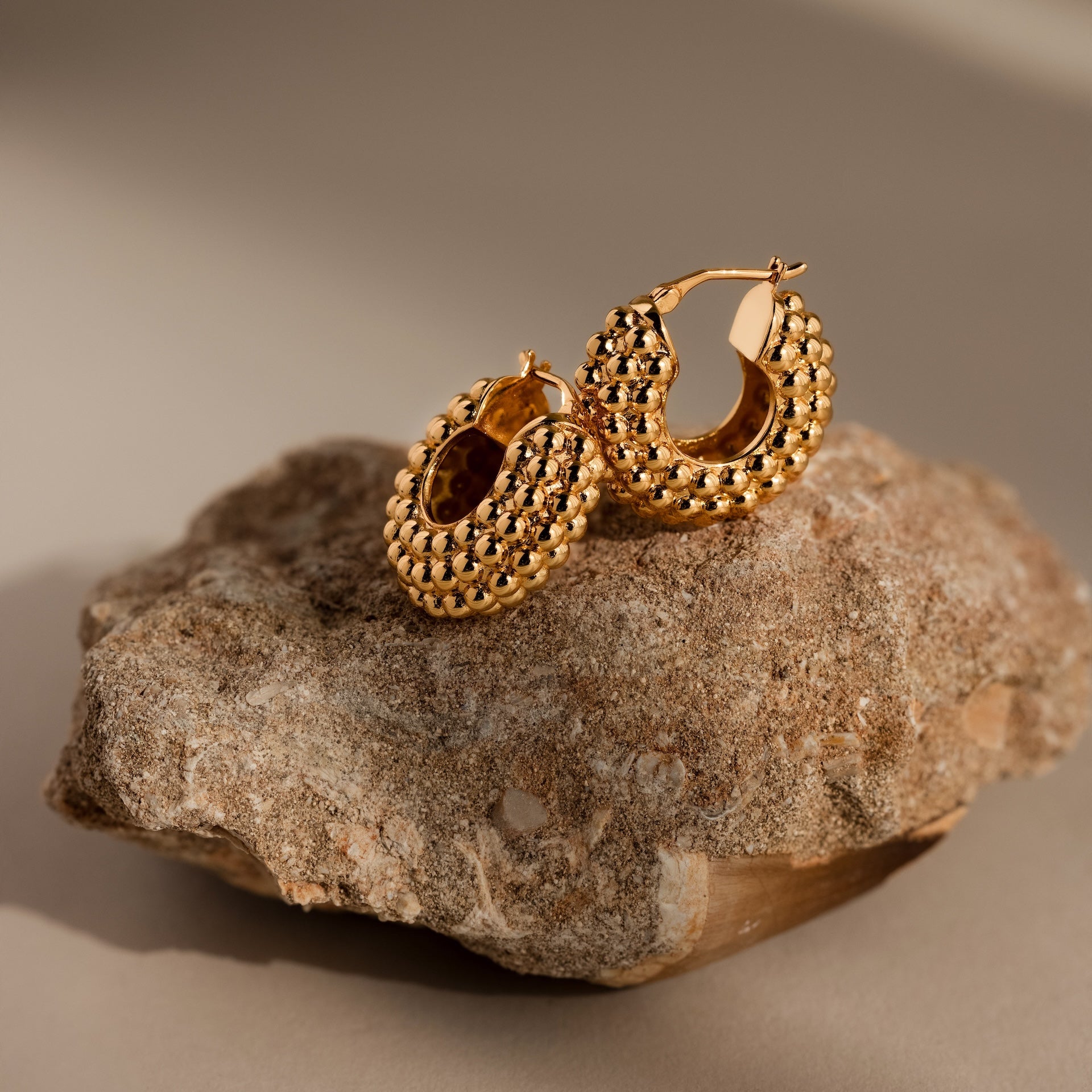Beaded Dome Hoops in 18K Gold are displayed on a rough stone surface, their bold hoop design standing out against a neutral background.