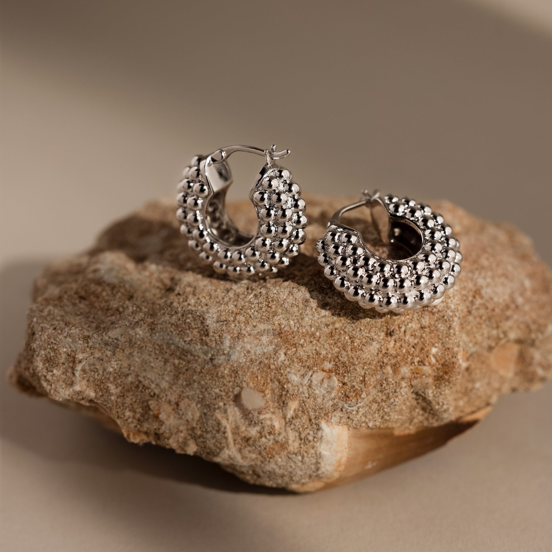 Beaded Dome Hoops, featuring delicate gold beading, are shown on a textured beige stone with a soft neutral background—ideal statement earrings for any occasion.