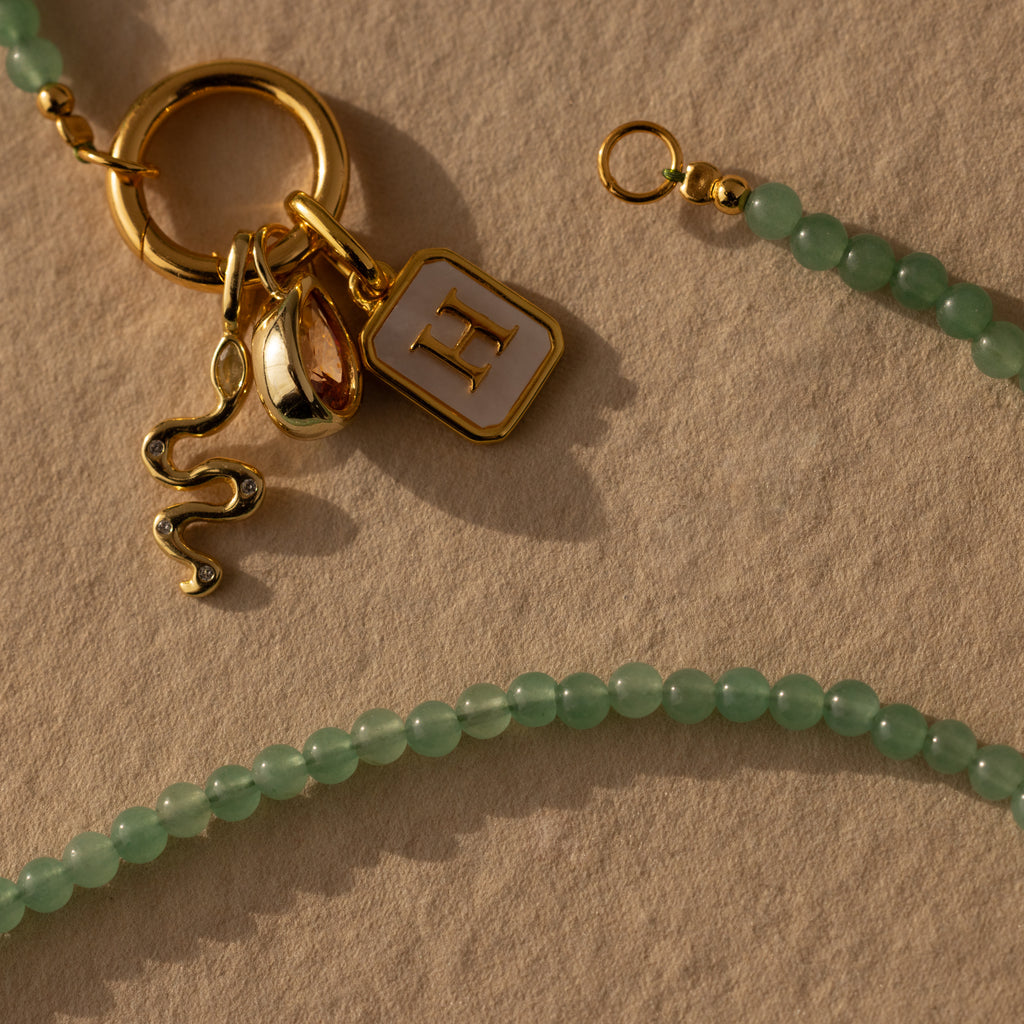 The Beaded Jade Clasp Necklace in 18K Gold features green jade beads, gold snake and letter H charms, and a classic gold clasp. Perfect for layering, it’s styled here on a textured beige surface.