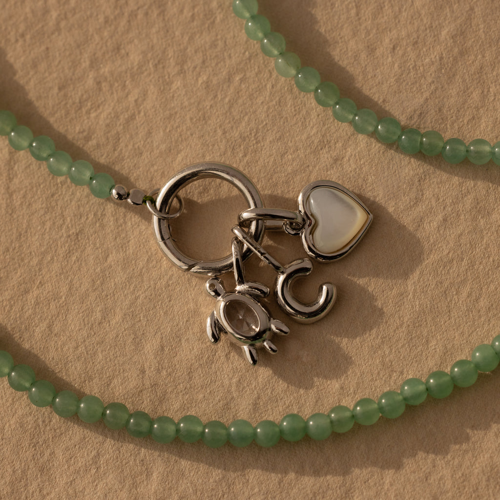 Beaded Jade Clasp Necklace in Silver features green jade beads, silver letter C, heart, and crab charms, plus a gold clasp—ideal for layering. Shown on a beige textured background.