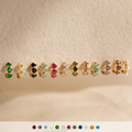 Nine pairs of gold earrings, including Triple Birthstone Huggies and minimalist hoops with colorful gemstones and pearls, are displayed on a textured beige surface.
