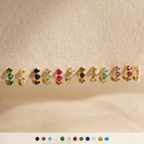 Nine pairs of gold earrings, including Triple Birthstone Huggies and minimalist hoops with colorful gemstones and pearls, are displayed on a textured beige surface.