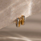 Dainty Mixed Metal Huggies, featuring gold and silver tones, are displayed on a textured light surface under soft lighting, emphasizing the elegance of minimalist jewelry.