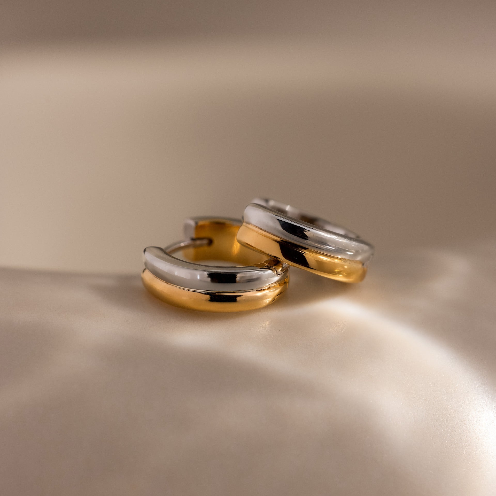 Dainty Mixed Metal Huggies in gold and silver are displayed on a smooth, cream-colored surface under soft lighting—ideal minimalist earrings for lovers of elegant two-tone jewelry.