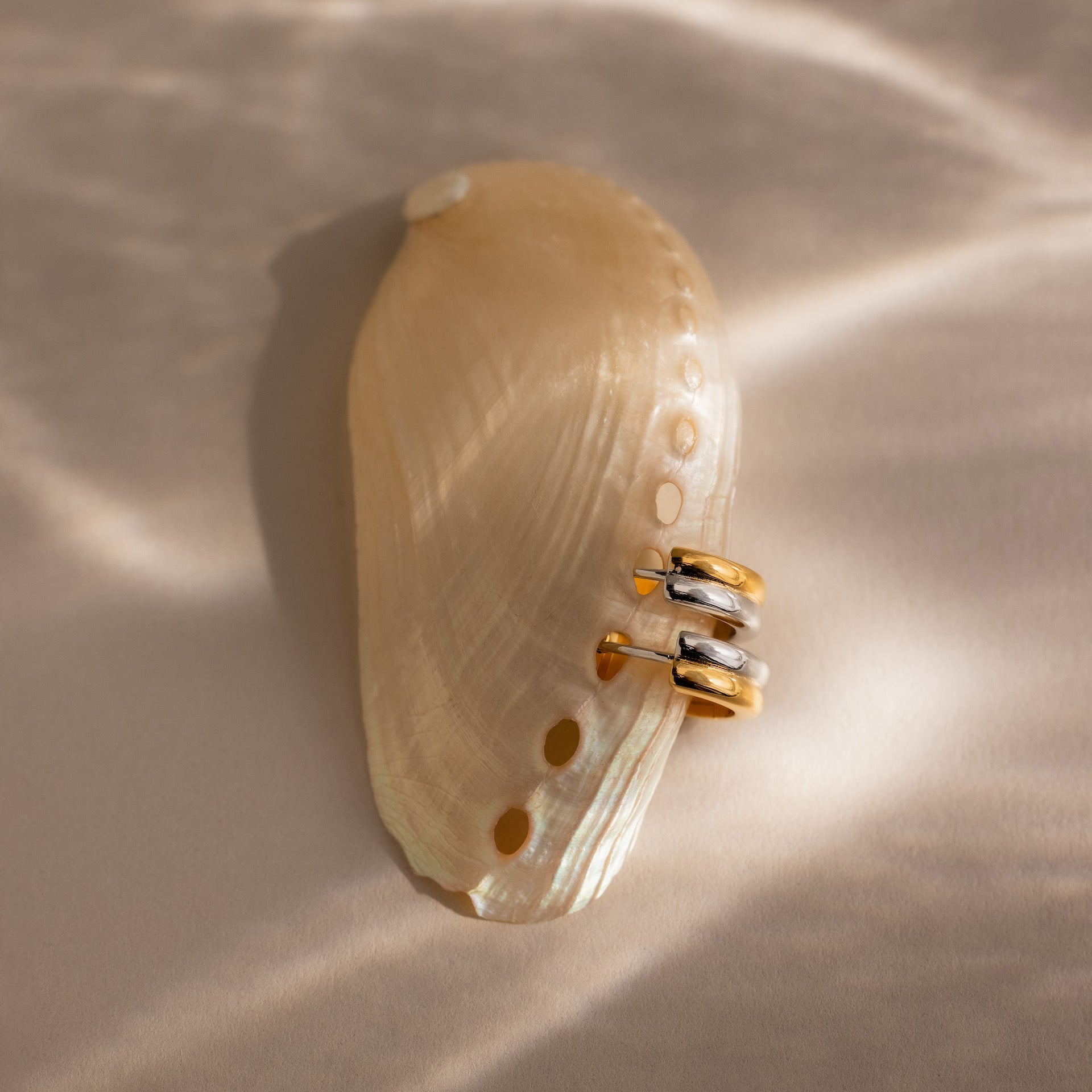 Dainty Mixed Metal Huggies in gold and silver rest on a beige seashell, aglow with soft, wavy light—a perfect minimalist accent for jewelry lovers.