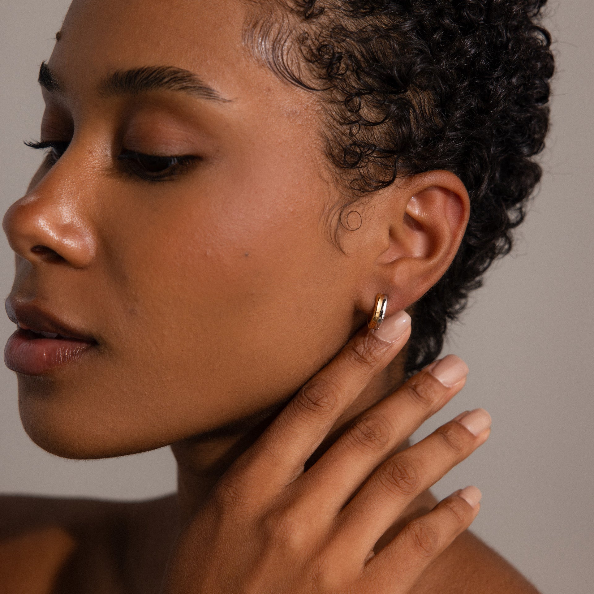 A woman with short curly hair touches her Dainty Mixed Metal Huggies, highlighting smooth skin and natural makeup—an ideal showcase of minimalist jewelry.
