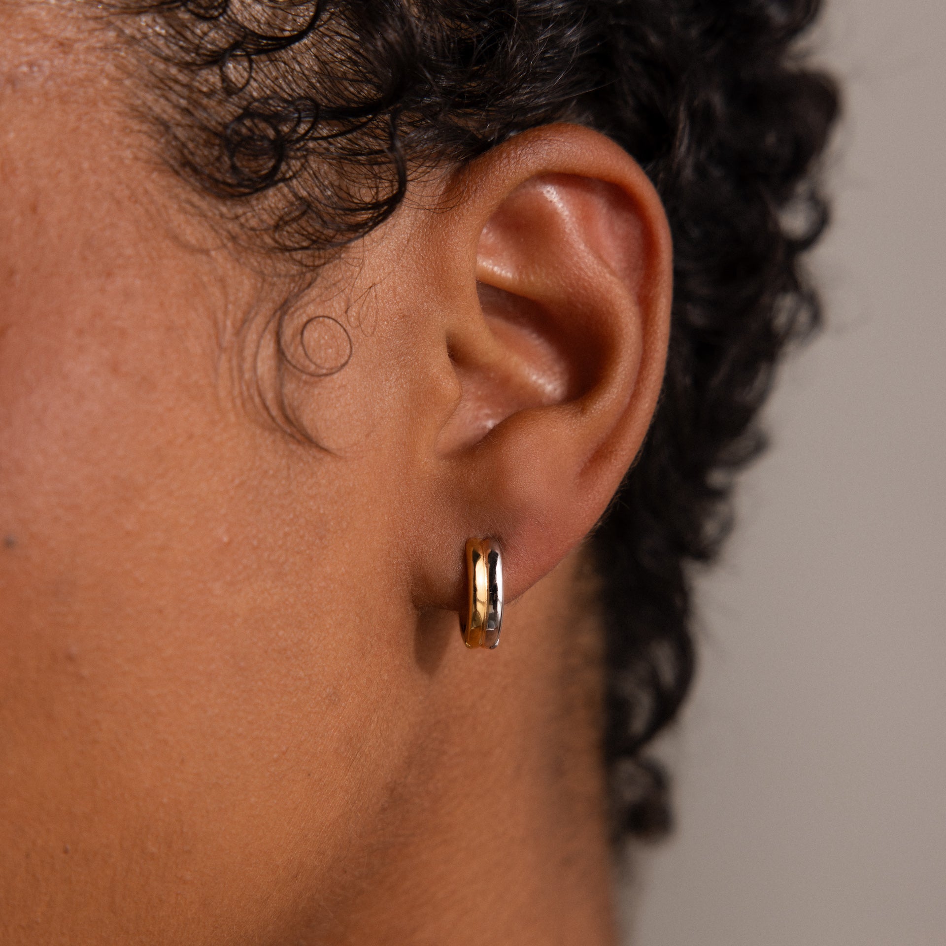 Close-up of a person’s ear wearing Dainty Mixed Metal Huggies with short curly hair, showcasing minimalist jewelry perfect for mixing and matching with gold and silver earrings.