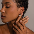 A woman with smooth skin wears Bold Mixed Metal Huggies, touching her ear with manicured fingers against a neutral background.