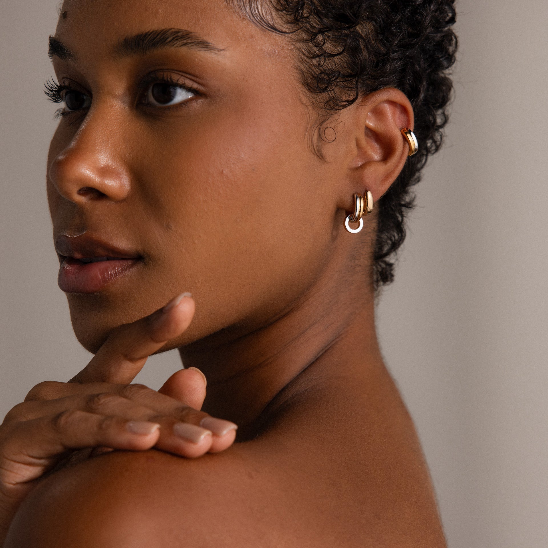 A woman with short curly hair touches her chin and gazes to the side, wearing Mixed Metal Circle Hoops in gold and silver.