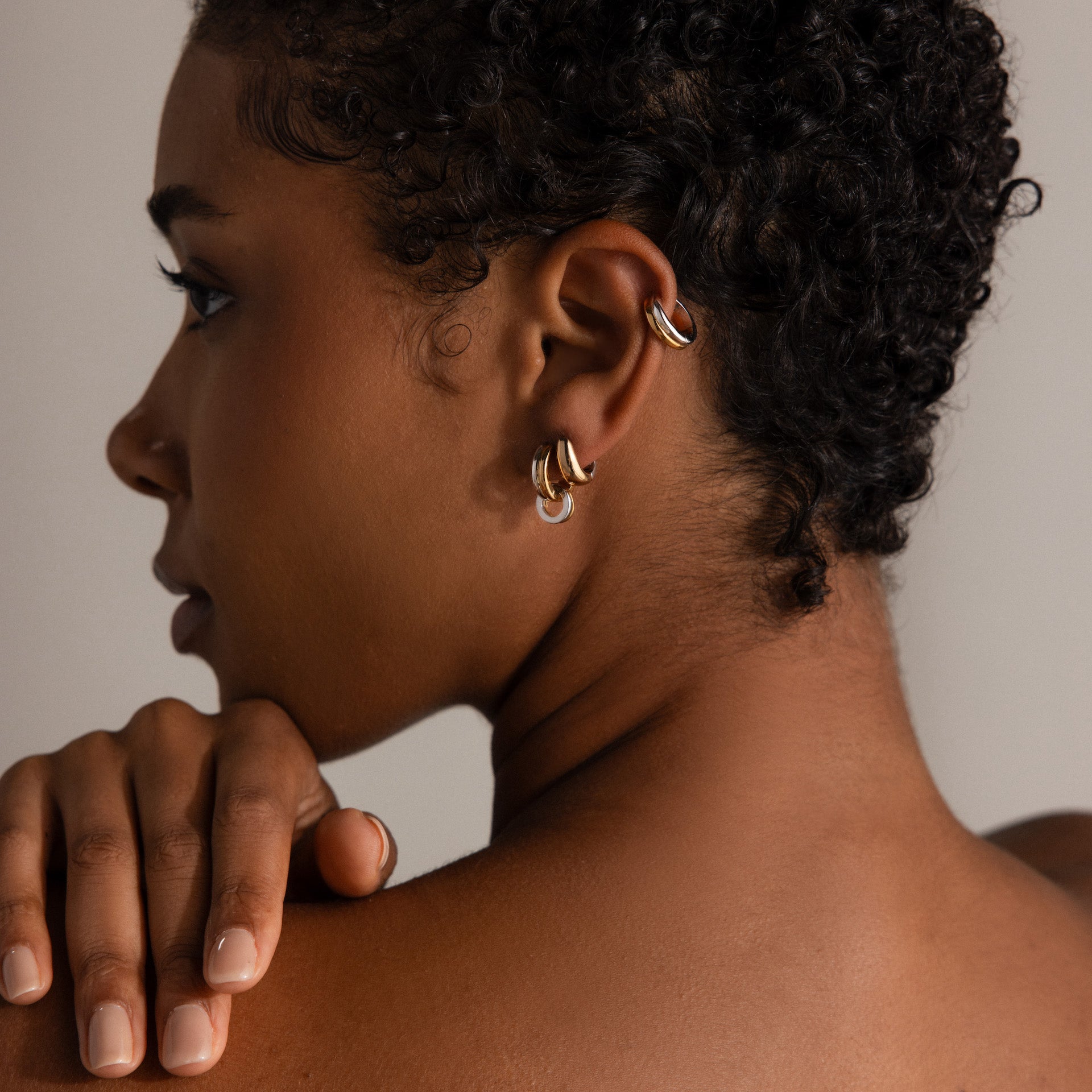 A woman with short curly hair wears minimalist jewelry—Dainty Mixed Metal Huggies earrings and a cuff—as she rests her hand on her shoulder.