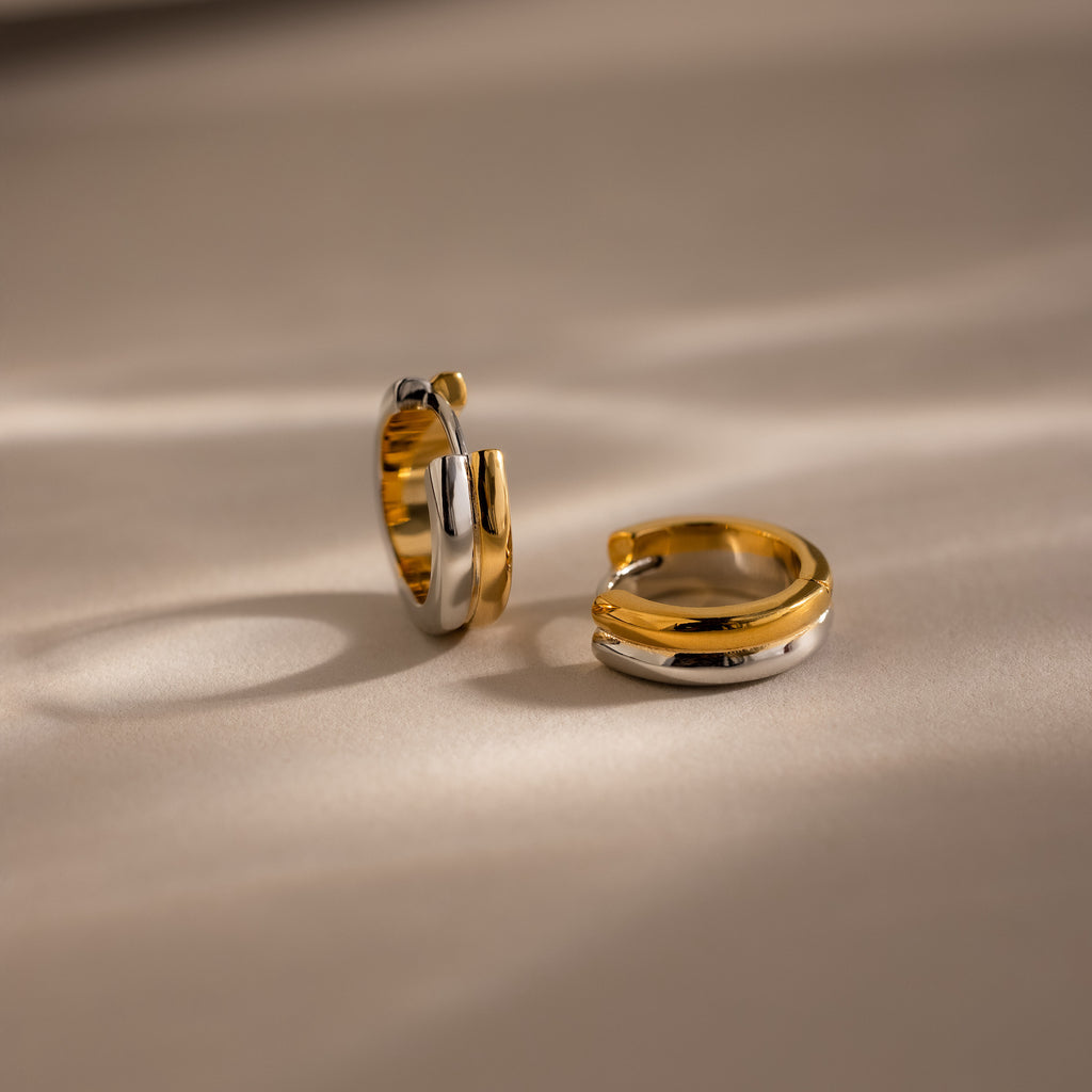The Dainty Mixed Metal Huggies in gold and silver rest on a beige surface with soft shadows, ideal for those who appreciate minimalist jewelry.