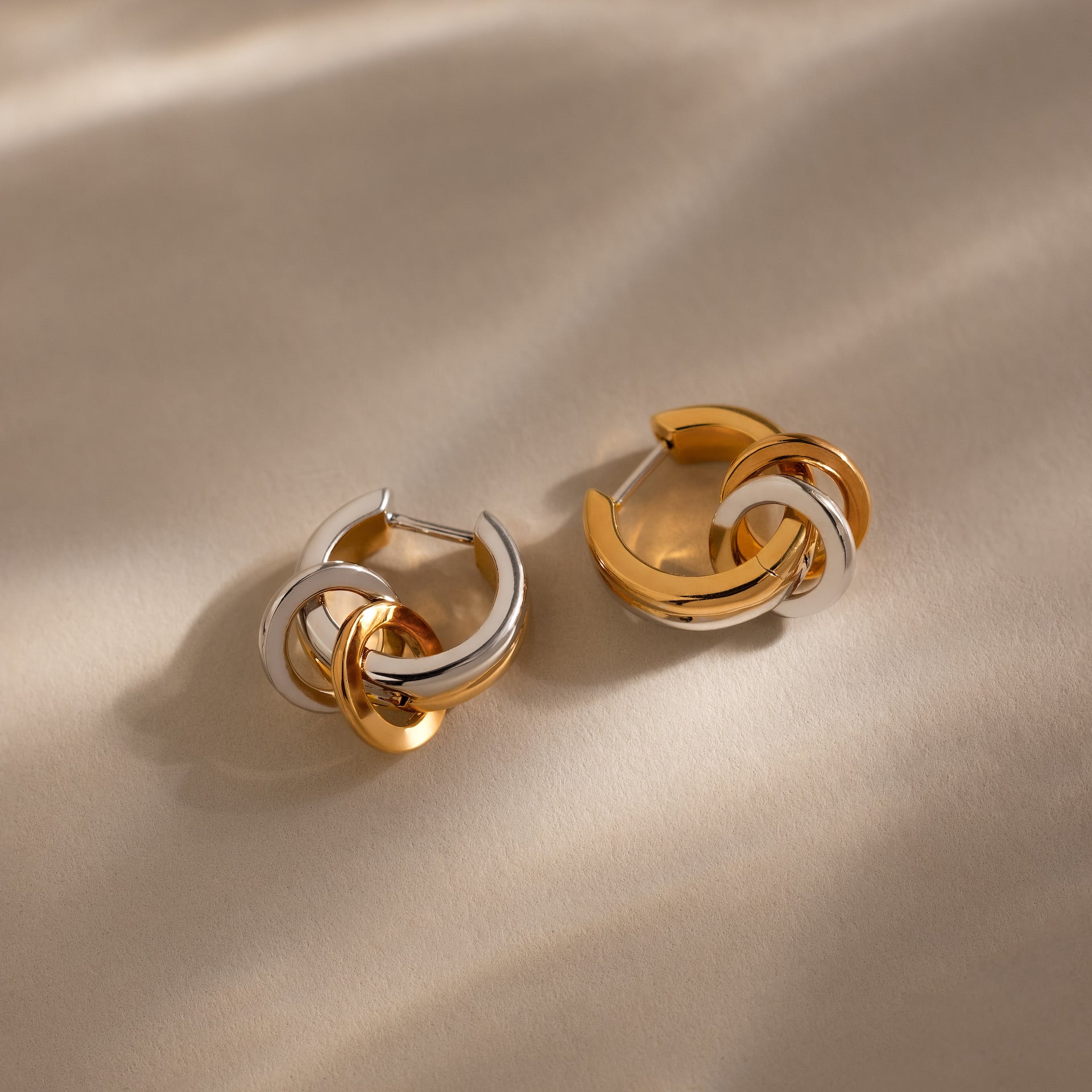 The Mixed Metal Circle Hoops, featuring intertwined gold and silver tones, rest on a neutral beige surface—versatile earrings perfect for any occasion.