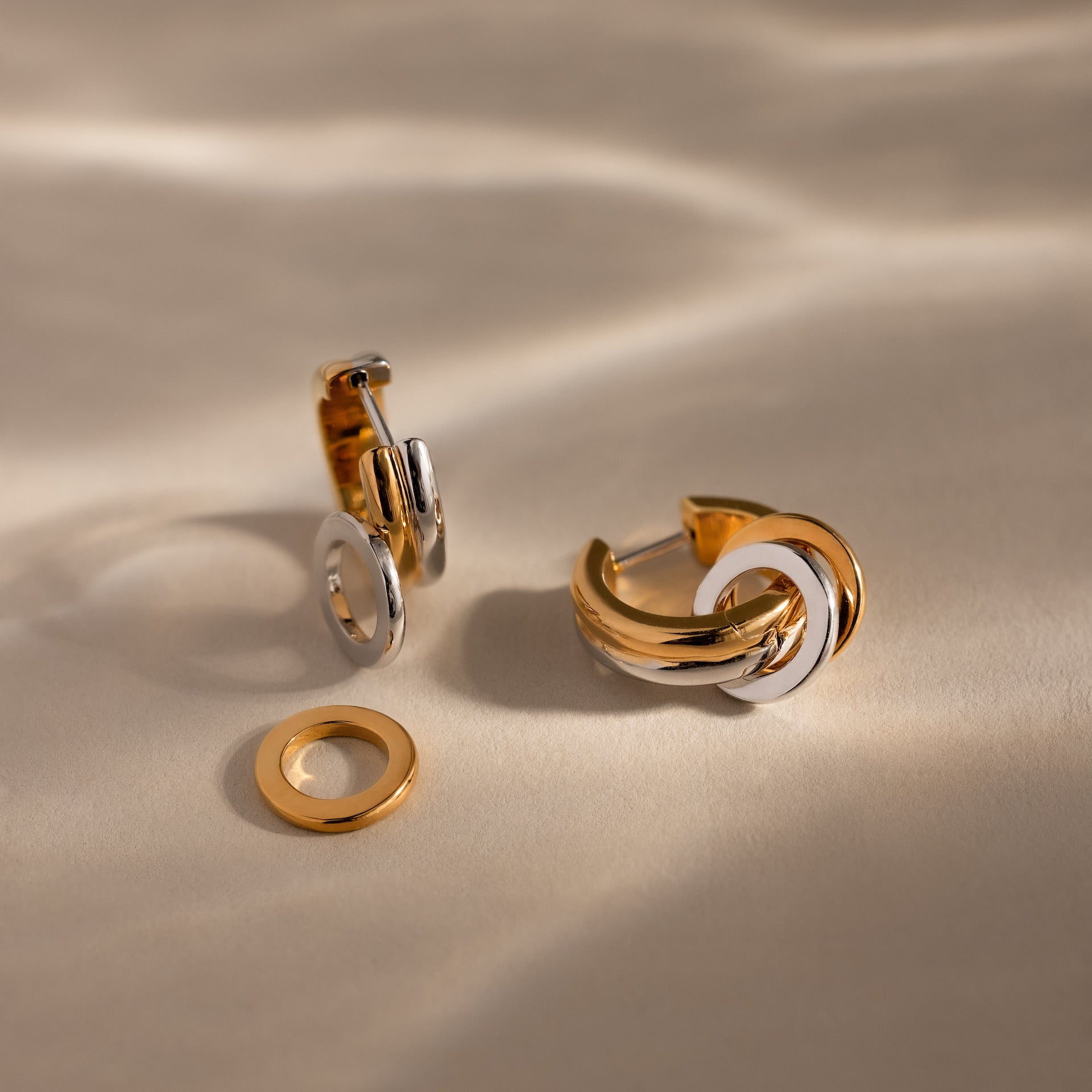 The Mixed Metal Circle Hoops combine gold and silver in two elegant earrings with matching circular charms, shown on a beige surface.