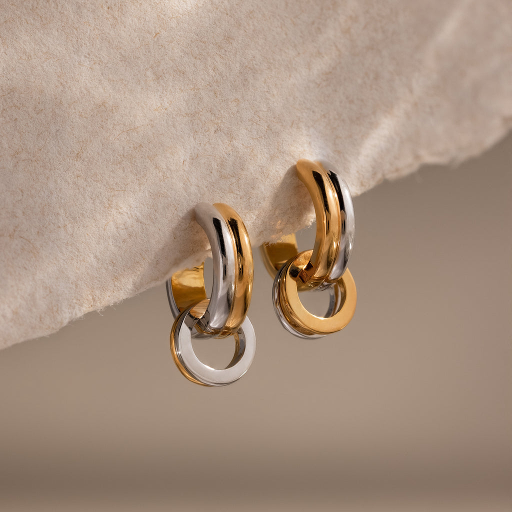 The Mixed Metal Circle Hoops feature interlocking gold and silver circles, elegantly displayed on a textured beige surface—a stylish example of versatile jewelry.