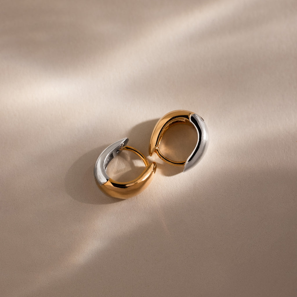 Bold Mixed Metal Huggies—one gold, one silver—sit on a beige surface with soft shadows, capturing the stylish, modern flair of these statement earrings.