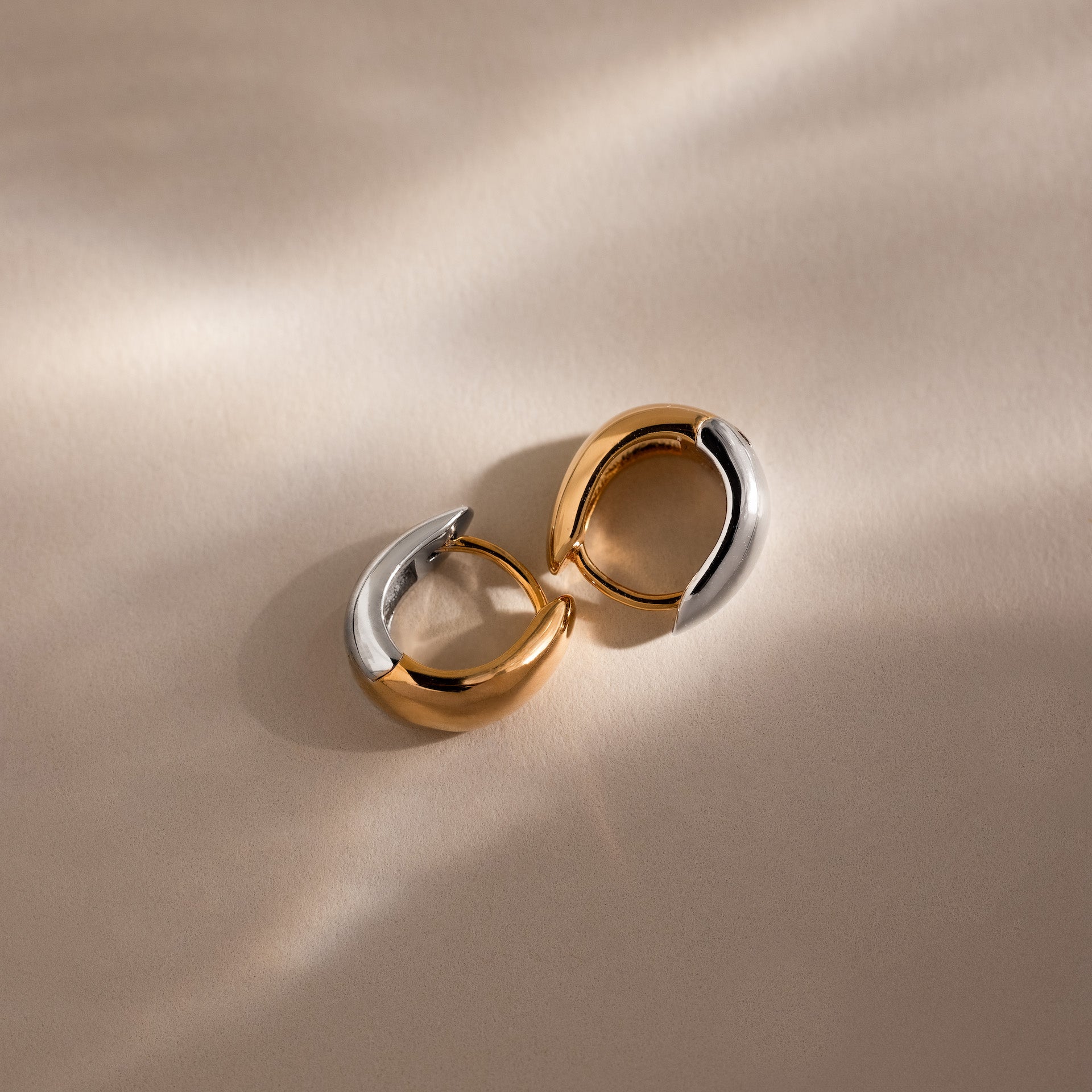 Bold Mixed Metal Huggies—one gold, one silver—sit on a beige surface with soft shadows, capturing the stylish, modern flair of these statement earrings.