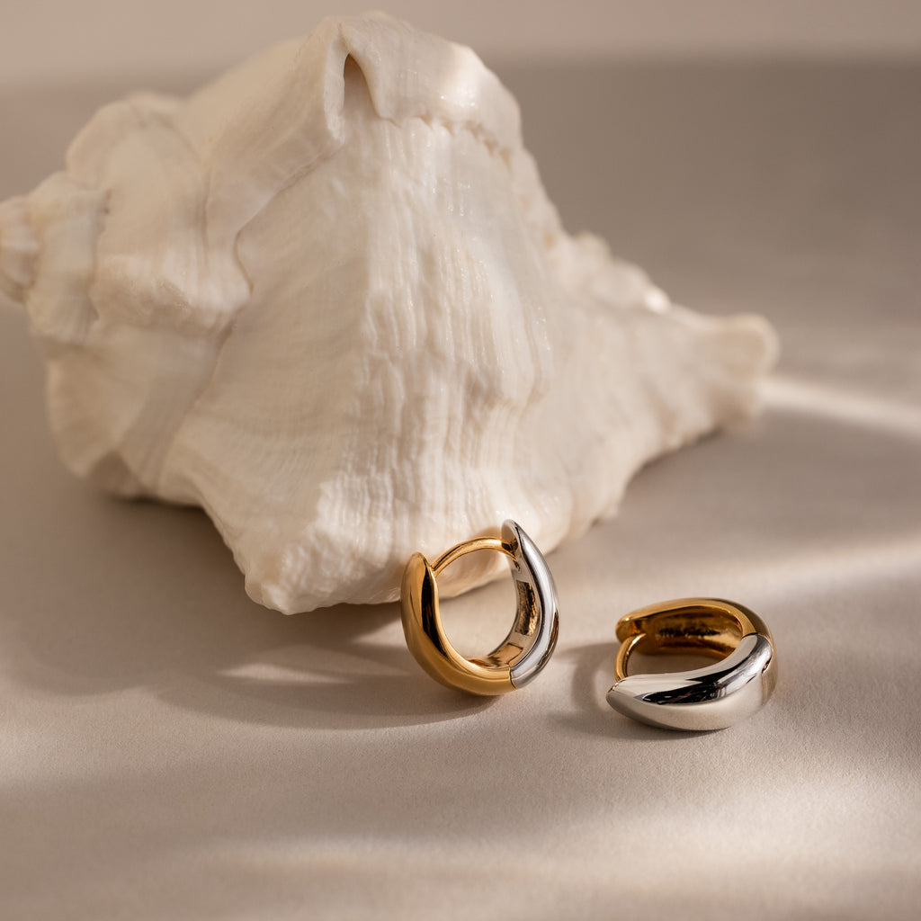 The Bold Mixed Metal Huggies—one gold and one silver—are displayed before a large white seashell, offering a bold, modern statement with these chunky contemporary earrings.