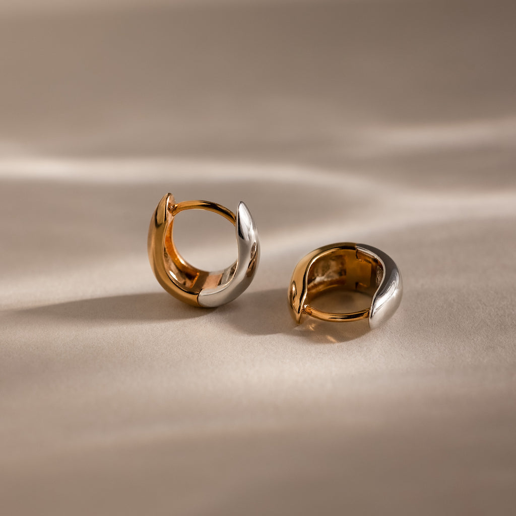 The Bold Mixed Metal Huggies—striking statement earrings in gold and silver—are displayed on a smooth beige surface, aglow in soft natural light.