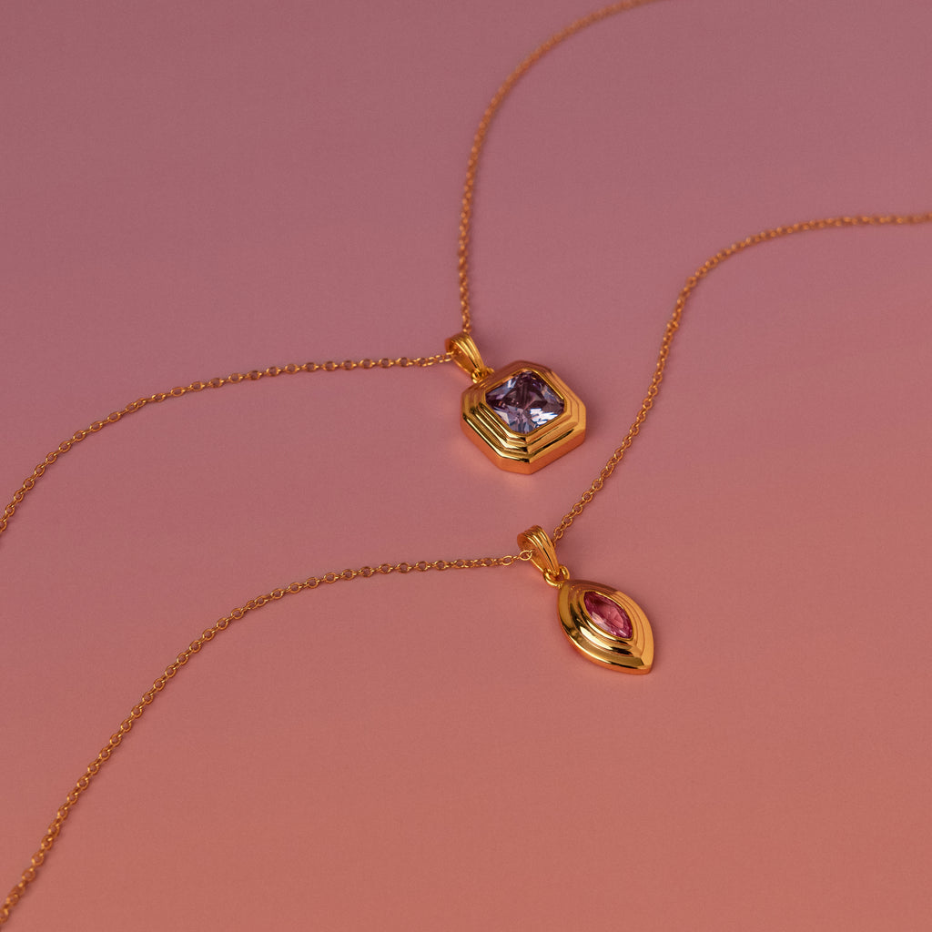 Two Ribbed Alexandrite Pendant Necklaces, each with a vibrant gemstone pendant in purple and pink, are displayed elegantly on a gradient pink background.