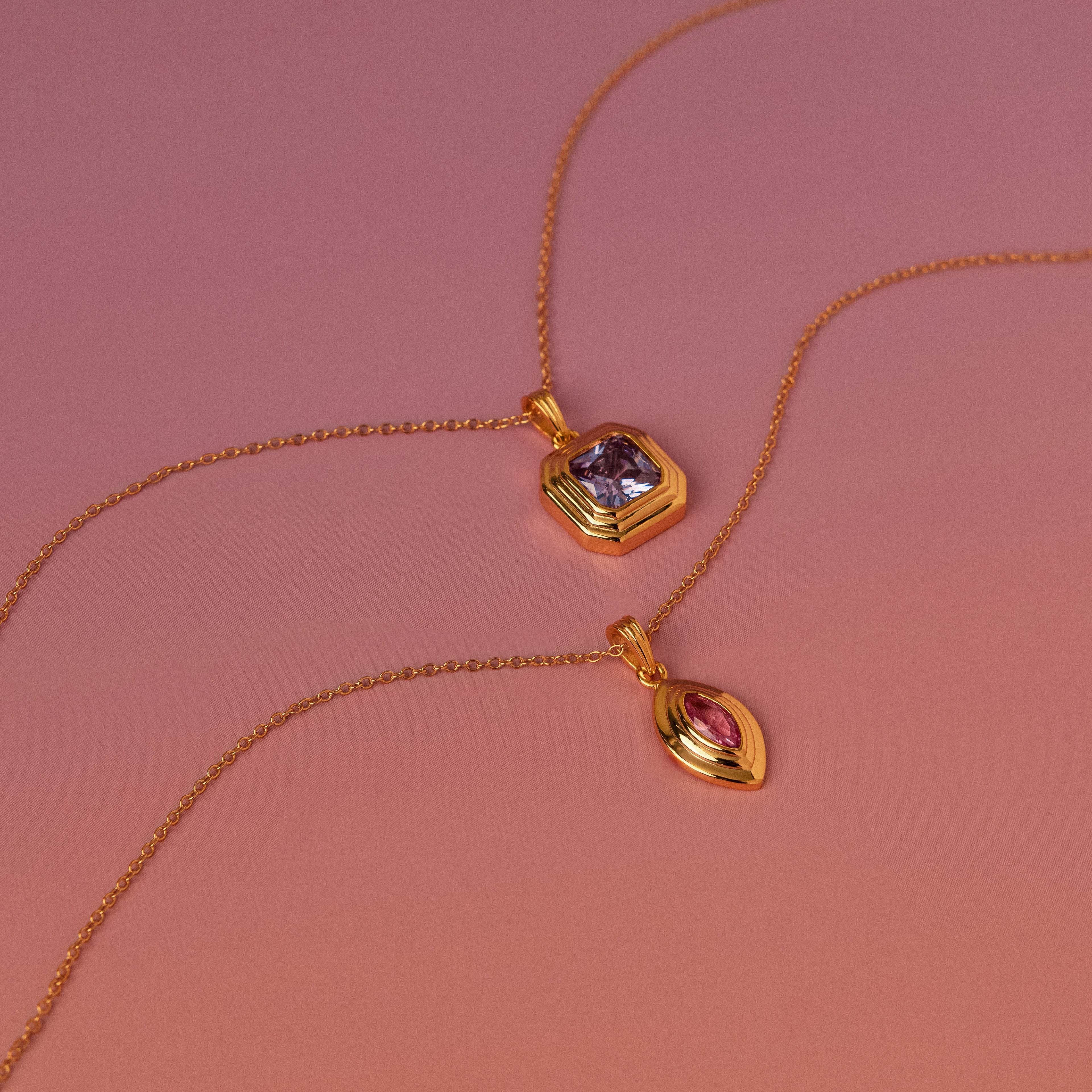 Two Ribbed Alexandrite Pendant Necklaces, each with a vibrant gemstone pendant in purple and pink, are displayed elegantly on a gradient pink background.