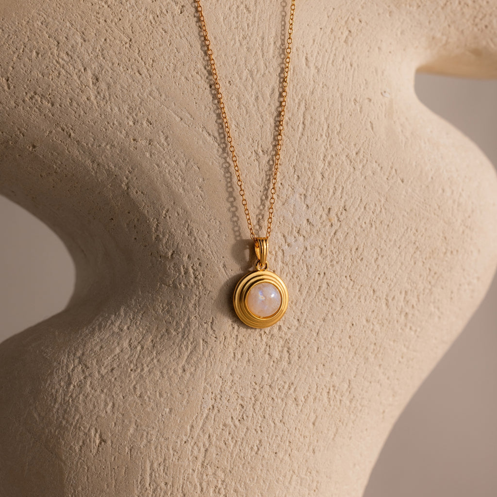 Ribbed Moonstone Pendant Necklace featuring a gold ribbed chain and a round white moonstone pendant, displayed on a textured beige sculpture.